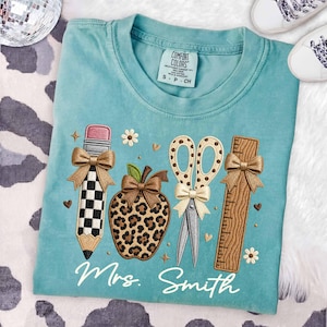 May include: A teal t-shirt featuring an embroidered design of a checkered pencil, a leopard print apple, a pair of scissors, and a wooden ruler, each accented with small bows. The text Mrs. Smith is embroidered in white script below the items.