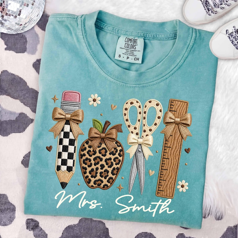 May include: A teal t-shirt featuring an embroidered design of a checkered pencil, a leopard print apple, a pair of scissors, and a wooden ruler, each accented with small bows. The text Mrs. Smith is embroidered in white script below the items.