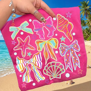 Può includere: Una maglietta rosa acceso con illustrazioni colorate di fiocchi, conchiglie e stelle marine. I fiocchi presentano vari motivi e colori, tra cui blu, arancione e viola. Perle bianche e stelle circondano i disegni. Lo sfondo mostra una scena di spiaggia con palme.
