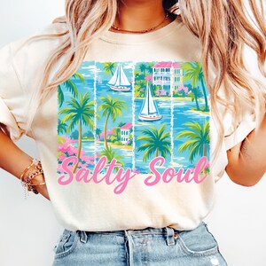 K&ouml;nnte beinhalten: Ein wei&szlig;es T-Shirt mit einem bunten Grafikdesign aus Palmen, Segelbooten und H&auml;usern. Der Text "Salty Soul" ist in Pink unterhalb der Grafik gedruckt.