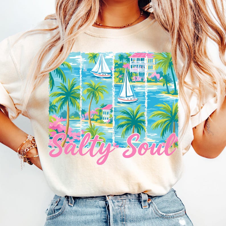 K&ouml;nnte beinhalten: Ein wei&szlig;es T-Shirt mit einem bunten Grafikdesign aus Palmen, Segelbooten und H&auml;usern. Der Text "Salty Soul" ist in Pink unterhalb der Grafik gedruckt.