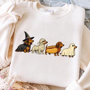 Peut inclure: Un sweat-shirt &agrave; col rond couleur cr&egrave;me avec une impression graphique de quatre teckels en costumes d'Halloween. Les chiens sont d&eacute;guis&eacute;s en sorci&egrave;re avec une cape et un chapeau noirs, en momie, en hot-dog et en fant&ocirc;me sous un drap blanc.