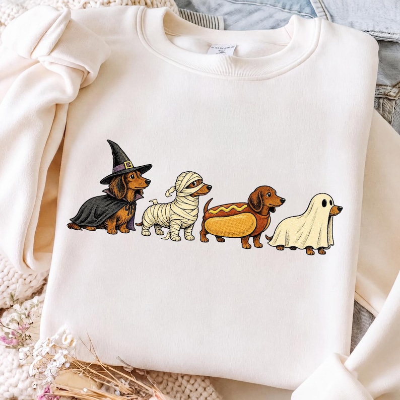 Peut inclure: Un sweat-shirt &agrave; col rond couleur cr&egrave;me avec une impression graphique de quatre teckels en costumes d'Halloween. Les chiens sont d&eacute;guis&eacute;s en sorci&egrave;re avec une cape et un chapeau noirs, en momie, en hot-dog et en fant&ocirc;me sous un drap blanc.