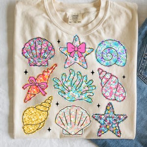 May include: A beige t-shirt with a floral print of seashells, starfish, and coral. The text "COMFORT COLORS XL - TO - EO" is printed on the shirt.