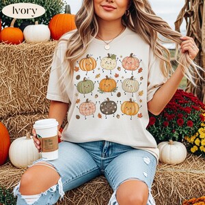 Puede incluir: Una camiseta de manga corta color marfil con un estampado gr&aacute;fico de nueve calabazas estilizadas con caras, brazos y piernas. Las calabazas est&aacute;n coloreadas en tonos naranja, verde, rosa y marr&oacute;n, rodeadas de peque&ntilde;as hojas de oto&ntilde;o y bellotas.
