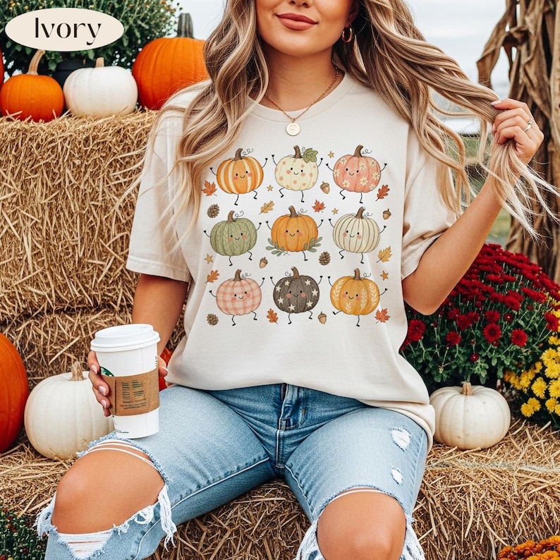 Puede incluir: Una camiseta de manga corta color marfil con un estampado gr&aacute;fico de nueve calabazas estilizadas con caras, brazos y piernas. Las calabazas est&aacute;n coloreadas en tonos naranja, verde, rosa y marr&oacute;n, rodeadas de peque&ntilde;as hojas de oto&ntilde;o y bellotas.
