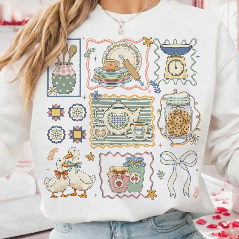 May include: White sweatshirt featuring a collage of kitchen-themed illustrations. The design includes images of a teapot, plates, a scale, cookies in a jar, ducks, and jars of jam. The illustrations are framed with decorative borders and floral accents.