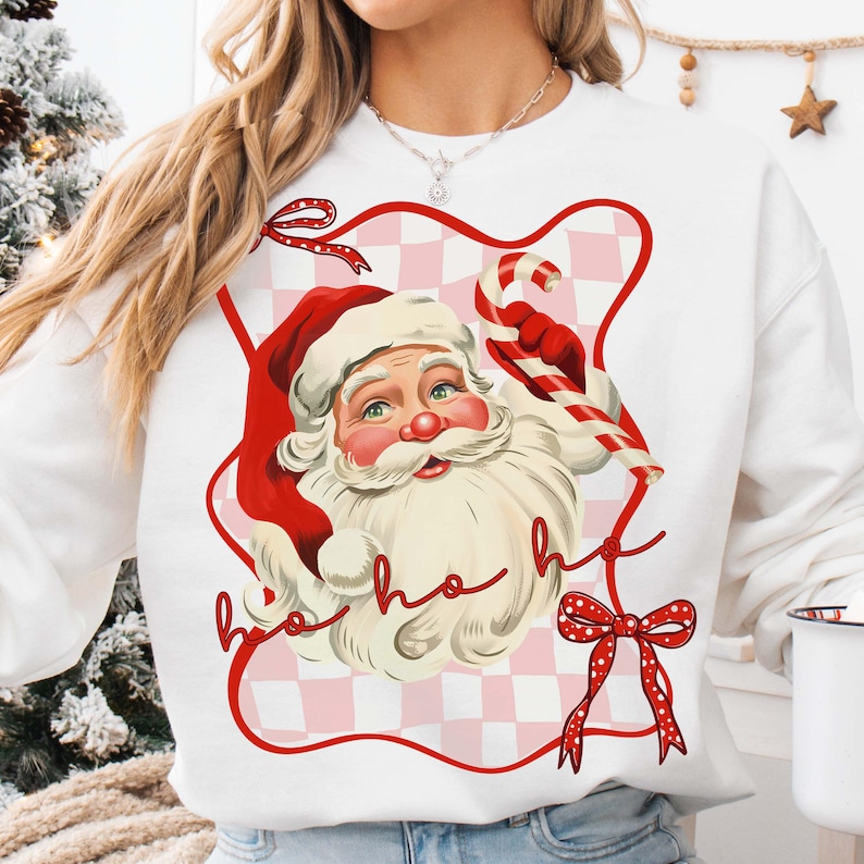 May include: White sweatshirt featuring a vintage illustration of Santa Claus with a red hat and beard, holding a candy cane. The design includes a red and pink checkered background and the words "ho ho ho".