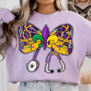 Puede incluir: Sudadera morada con un diseño de lazo temático de Mardi Gras con una flor de lis y cuentas. El lazo está formado por dos estetoscopios.