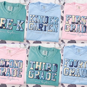 May include: Six colorful t-shirts with grade level text. The shirts are light blue, pink, and teal. Each shirt has a grade level embroidered in a patchwork style with floral accents. The text includes "PRE-K", "Kindergarten", "First Grade", "Second Grade", "Third Grade", and "Fourth Grade".