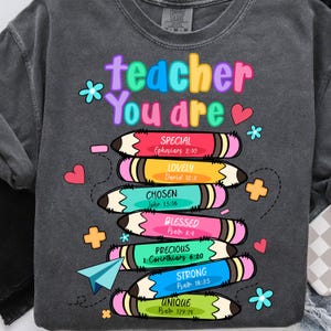 May include: Dark gray sweatshirt with the words "teacher You are" in colorful letters. Below are stacked pencils with words like "SPECIAL", "LOVELY", and "CHOSEN". The sweatshirt has a crew neck and long sleeves. Floral and heart graphics are scattered around the design.