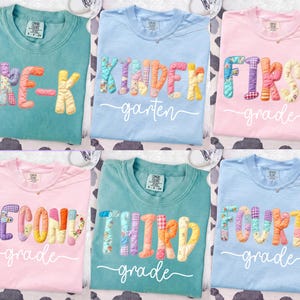 May include: Six colorful t-shirts with school grade level text. The shirts are light blue, pink, and teal. The text includes "Pre-K", "Kindergarten", "First Grade", "Second Grade", "Third Grade", and "Fourth Grade". The text is in a patchwork style.