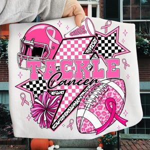 Può includere: Asciugamano bianco con un casco da football americano rosa, un pallone da football e un pompon. Le parole "TACKLE Cancer" sono in rosa. Il design include nastri rosa, stelle e un megafono con la parola "CHEER".