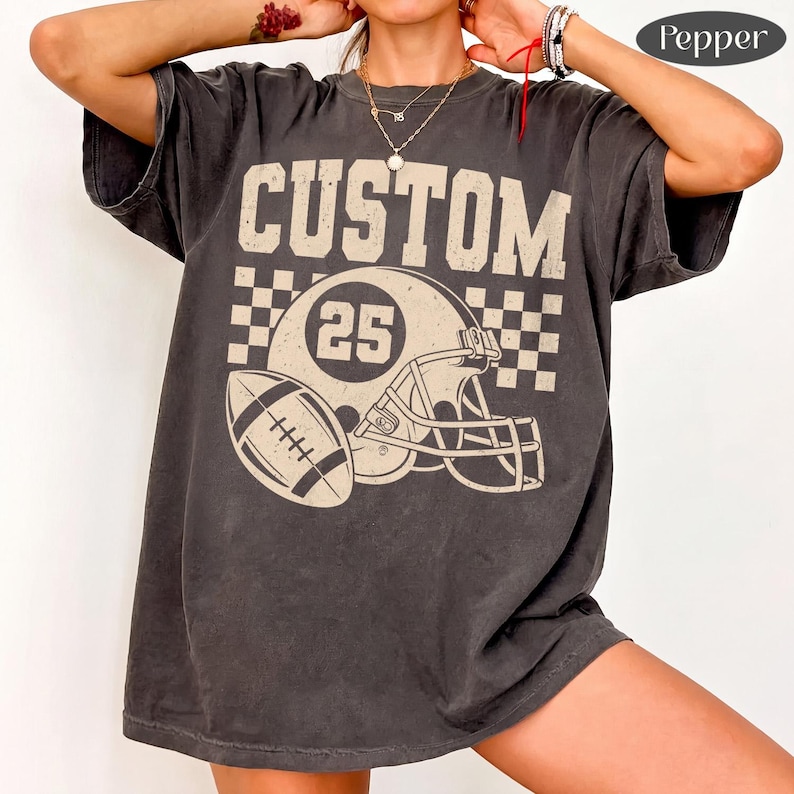 May include: A charcoal grey short-sleeve t-shirt featuring a distressed cream-colored graphic. The design includes the word CUSTOM above a football helmet with the number 25, a football, and a checkered pattern background.