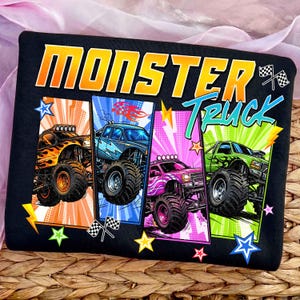 May include: Black t-shirt featuring the words "MONSTER TRUCK" in orange and teal. The shirt displays four colorful monster truck designs with flames, lightning bolts, and stars. Each truck is a different color: orange, blue, pink, and green.