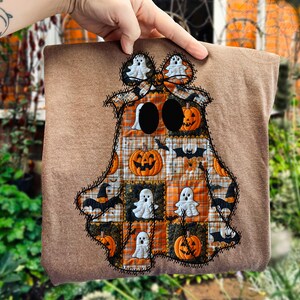 May include: A brown Halloween-themed square with a patchwork ghost design. The ghost is made of orange and brown plaid squares, with embroidered details, pumpkins, bats, and ghost figures. The edges are stitched with black thread.
