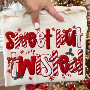 May include: Cream-colored sweatshirt with the phrase "Sweet but Twisted" in red and white, festive lettering. The design includes candy cane stripes, snowflakes, and holly accents, perfect for the holiday season.