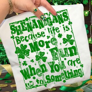 May include: White t-shirt with green text that reads "SHENANIGANS Because life is More FUN When You are up to Something." The design includes shamrocks and decorative elements, perfect for St. Patrick's Day.