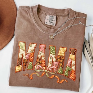 May include: A brown short-sleeve t-shirt featuring the word MAMA in large, patchwork-style letters with various floral, plaid, and leaf patterns in orange, green, and yellow. A decorative bow is embroidered below the text, and a silver necklace is visible.