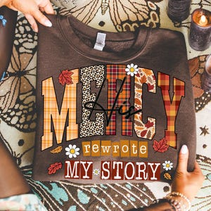 May include: A brown sweatshirt with the words "MERCY His rewrote MY STORY" in a mix of plaid, leopard print, and floral patterns. The design includes fall leaves and daisy accents. The sweatshirt is held up by a person.