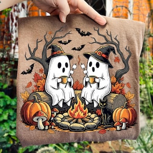 May include: A brown t-shirt featuring a Halloween-themed graphic. Two white ghosts wearing witch hats are seated by a campfire, holding mugs and marshmallows. Pumpkins, fall leaves, bats, and a black cat complete the design.