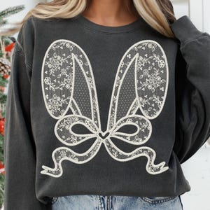 Op de afbeelding: Een donkergrijs sweatshirt met een wit kantontwerp van konijnenoren en een strik. Het ontwerp is gecentreerd op de voorkant van het sweatshirt.