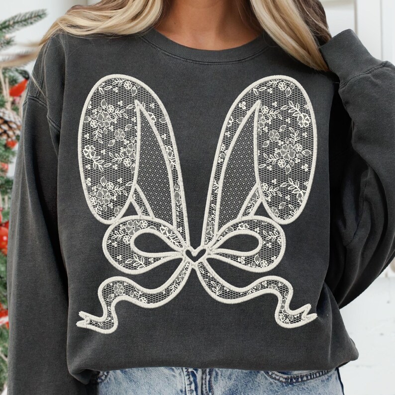 Op de afbeelding: Een donkergrijs sweatshirt met een wit kantontwerp van konijnenoren en een strik. Het ontwerp is gecentreerd op de voorkant van het sweatshirt.