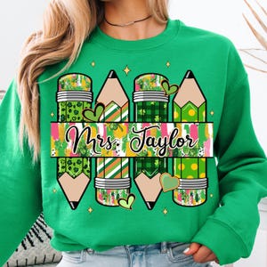 May include: Green sweatshirt featuring a pencil design with shamrocks and the text "Mrs. Taylor". The pencils have various patterns, including polka dots and plaid. The design includes green hearts and gold stars.