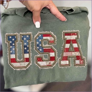 May include: An olive green sweatshirt with the word "USA" embroidered on the front. The letters are designed with a patchwork pattern in red, white, and blue, resembling the American flag. The embroidery has a frayed edge.