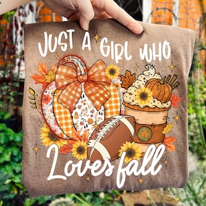 Puede incluir: Camiseta marrón con el texto "Just a Girl Who Loves Fall". El diseño incluye una calabaza, un balón de fútbol americano, una taza de café, girasoles y hojas de otoño. La calabaza tiene un lazo a cuadros.