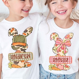 以下が含まれることがあります： イースターバニーのデザインが施された白いTシャツ2枚。1枚のシャツには、迷彩柄のウサギと「Andrew」という名前がバナーに描かれています。もう1枚のシャツには、ピンクのリボンと「Amelia」という名前が付いた花のウサギのデザインが施されています。どちらのシャツにもニンジンのグラフィックがあります。