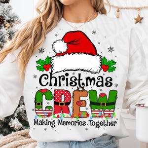 May include: White sweatshirt with a Christmas Crew design. The design includes a red Santa hat, holly, snowflakes, and the words "Christmas Crew" in colourful, patterned letters. The phrase "Making Memories Together" is below.