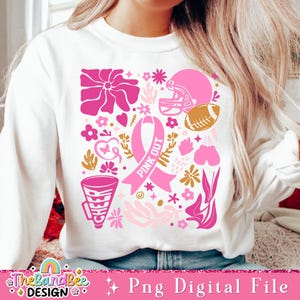 May include: A white sweatshirt with a pink and gold design featuring a pink ribbon, a football helmet, a football, and flowers. The ribbon has the text "Pink Out" on it.
