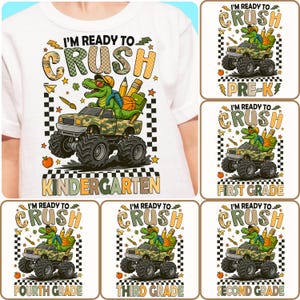 Pode incluir: Camiseta branca com um gráfico colorido. O design apresenta um dinossauro dirigindo um caminhão monstro camuflado com o texto "I'M READY TO CRUSH" e "KINDERGARTEN". Designs adicionais incluem "PRE-K", "FIRST GRADE", "FOURTH GRADE", "THIRD GRADE" e "SECOND GRADE".