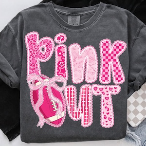 May include: Dark gray sweatshirt with the words "PINK OUT" in pink, with various patterns and a football graphic. The letters are decorated with leopard print, stars, hearts, and a checkered pattern. A pink bow is on the football.