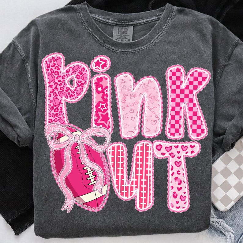 May include: Dark gray sweatshirt with the words "PINK OUT" in pink, with various patterns and a football graphic. The letters are decorated with leopard print, stars, hearts, and a checkered pattern. A pink bow is on the football.