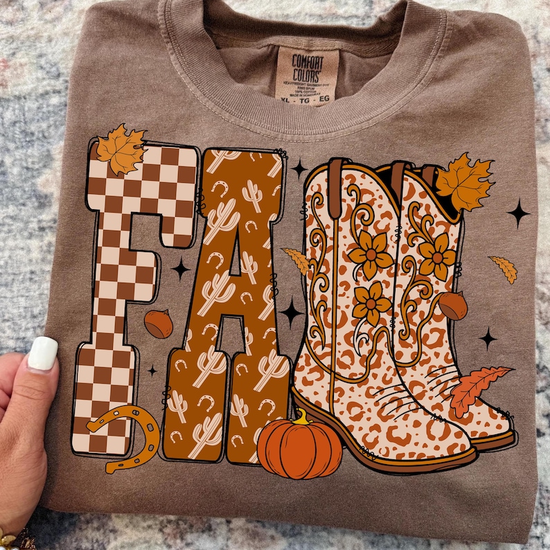 May include: Brown long-sleeved top featuring a fall design. The word "FALL" is displayed with a checkered pattern, cacti, and cowboy boots. A pumpkin, leaves, and a horseshoe complete the design.