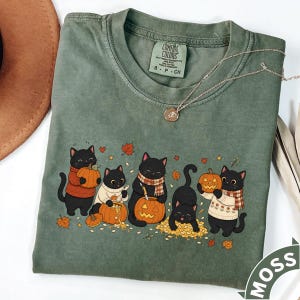 May include: A moss green crew neck t-shirt featuring a graphic of five black cats carving and playing with orange pumpkins. The cats wear sweaters and scarves, surrounded by autumn leaves. The shirt has a Comfort Colors brand label at the neck.
