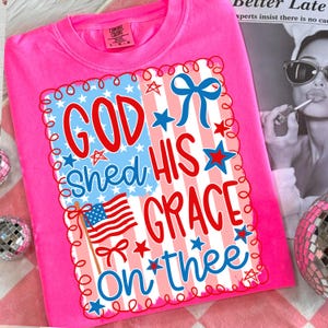 May include: A bright pink t-shirt with a patriotic design. The design features the text "GOD SHED HIS GRACE ON THEE" in red and blue, along with an American flag and stars. The shirt is made of a soft material.
