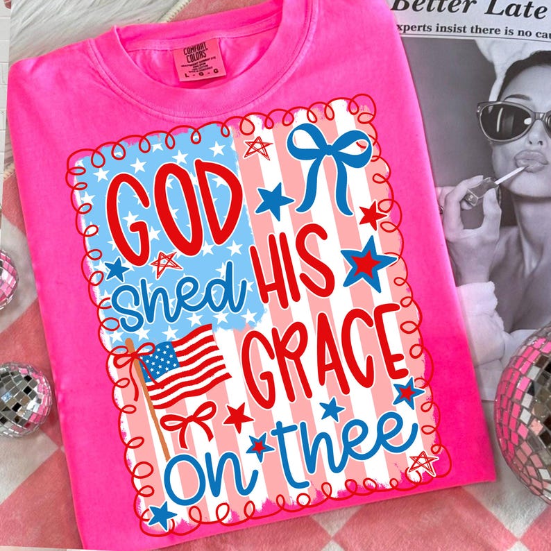 May include: A bright pink t-shirt with a patriotic design. The design features the text "GOD SHED HIS GRACE ON THEE" in red and blue, along with an American flag and stars. The shirt is made of a soft material.