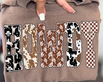 May include: A brown sweatshirt with the word "HOWDY" in a western-themed design. The letters feature cowhide, cactus, and checkered patterns. The letter "H" has cowboy-themed illustrations.