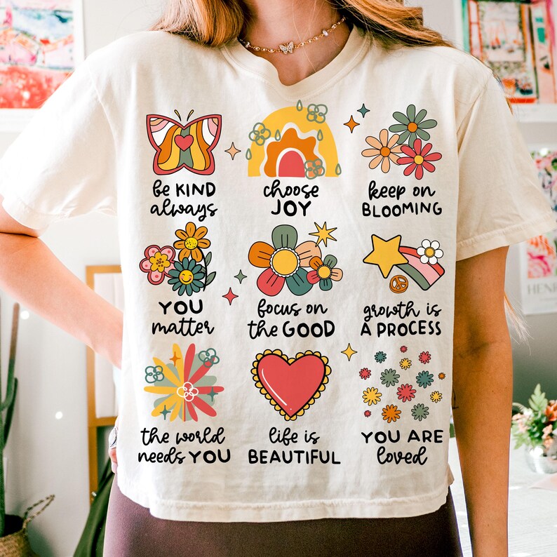 May include: A white t-shirt with a colorful graphic design featuring flowers, butterflies, and positive messages like "be kind always", "choose joy", "keep on blooming", "you matter", "focus on the good", "growth is a process", "the world needs you beautiful", and "you are loved".