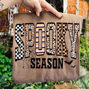 以下が含まれることがあります： 茶色のTシャツ。 「SPOOKY SEASON」の文字が、さまざまな模様と色で描かれています。 文字は市松模様、ヒョウ柄、オレンジ色のアクセントで装飾されています。 クモの巣とコウモリも描かれています。