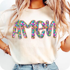 May include: A white t-shirt with the word "Amen" printed in large, colorful letters. The letters are filled with a floral pattern in shades of pink, blue, yellow, and green.