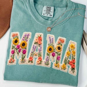May include: A seafoam green t-shirt with the word "MAMA" embroidered in colorful flowers. The letters are filled with sunflowers, tulips, roses, and lavender. A silver necklace with a small pendant rests on the shirt.