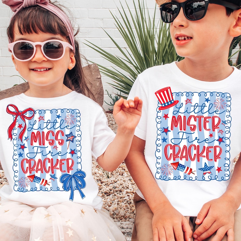 May include: Two children wearing white t-shirts with patriotic designs. The shirts feature the text "Little Miss Fire Cracker" and "Little Mister Fire Cracker" in red, white, and blue, with fireworks and star accents. The girl wears a pink headband and sunglasses.
