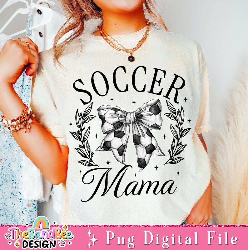 May include: A white t-shirt with black text that reads "Soccer Mama". The text is surrounded by a wreath of leaves and a black and white soccer ball bow.