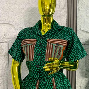 May include: A green and black patterned two-piece outfit on a mannequin. The short-sleeved top has two pockets with vertical stripes in brown, orange, and green. The outfit is buttoned up and has a matching skirt.