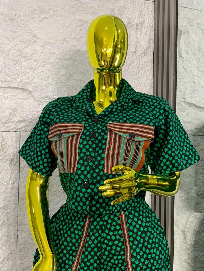 May include: A green and black patterned two-piece outfit on a mannequin. The short-sleeved top has two pockets with vertical stripes in brown, orange, and green. The outfit is buttoned up and has a matching skirt.