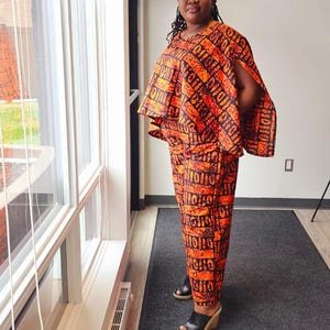 Puede incluir: Una mujer lleva un conjunto de dos piezas con una capa superior y pantalones. La tela es naranja y negra con un patrón geométrico. El conjunto tiene un diseño moderno y elegante.